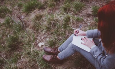Redhead girl sitting on green spring grass writing down her thoughts in notebook.