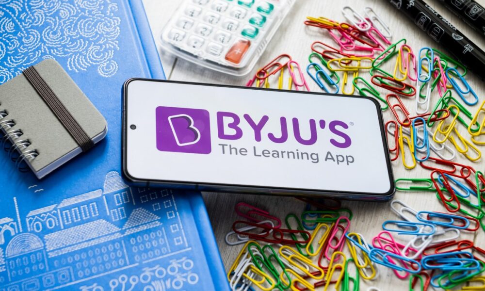 Byju's logo displayed on a smartphone laying on a table covered in school supplies